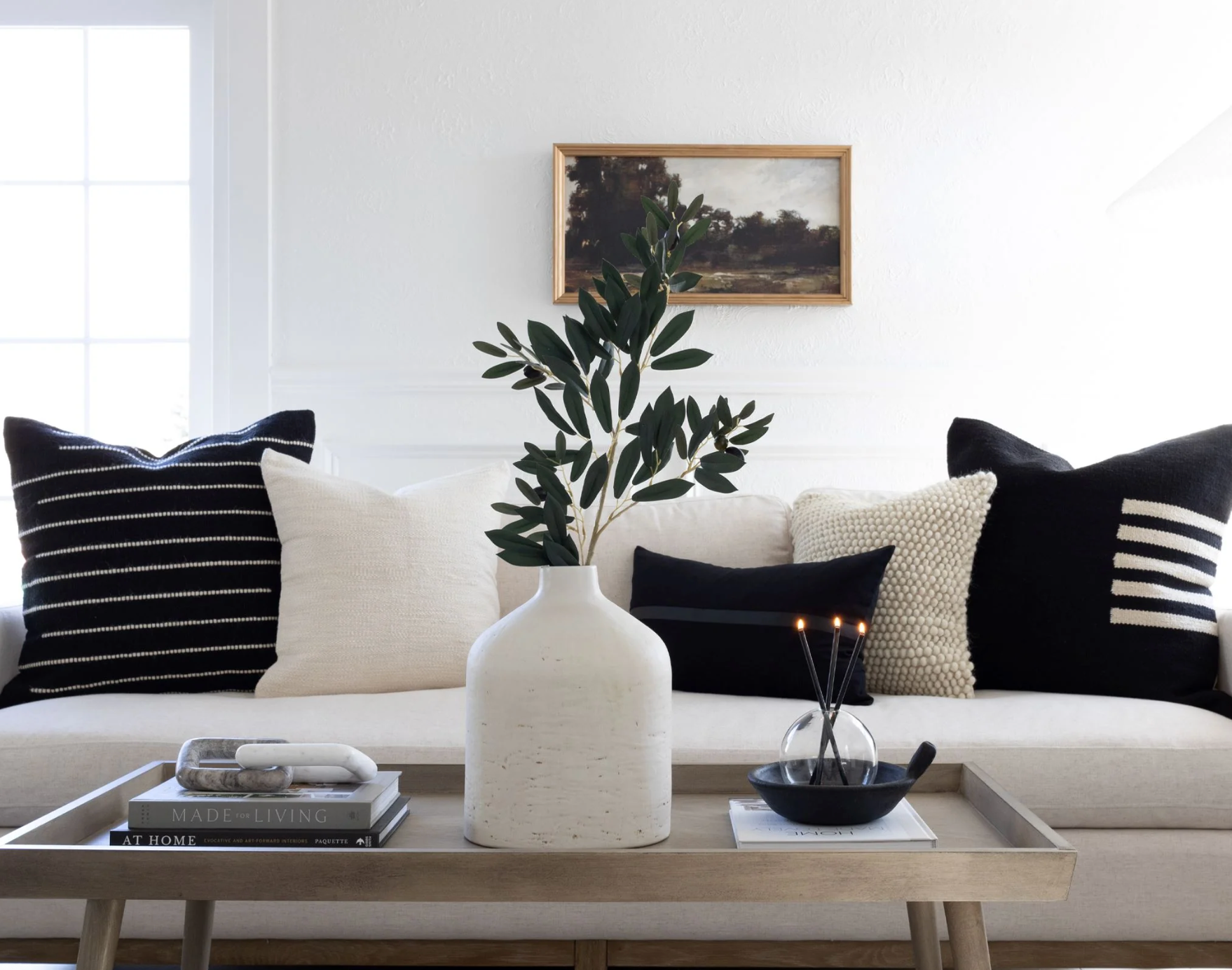 Black and cream pillow arrangement on a white linen sofa with layered textures — stripe, bobble, and solid pillow covers styled with an olive branch and vintage landscape art