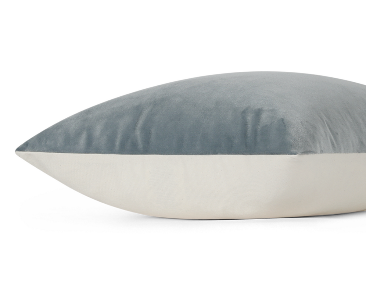 A close-up of a rectangular throw pillow with a slate blue velvet top half and a cream fabric bottom half, shown against a plain white background.