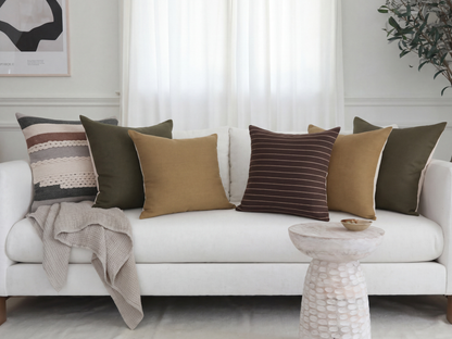 Crosby striped textured, olive linen, ochre linen, and umber striped wool pillow covers styled together on a cream sofa with a grey throw blanket