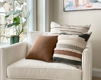 Sepia cotton pillow cover styled on a white armchair with stripe and textured cream pillow covers